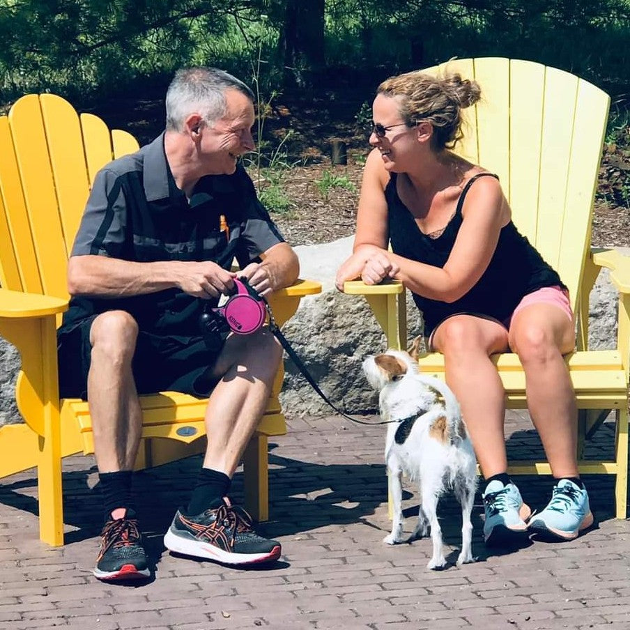 Two people sitting on yellow Adirondack chairs with a dog on a leash in a park setting.